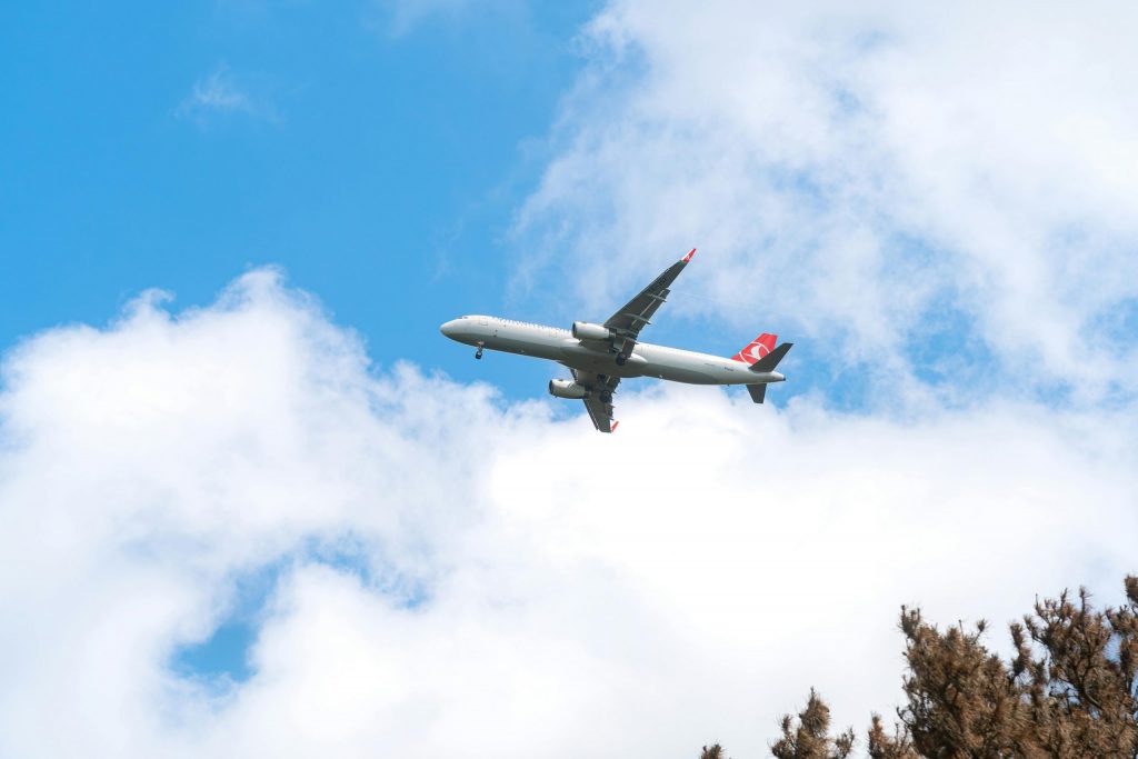 Plane flying through a blue sky with white clouds above treetops.
