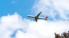 Plane flying through a blue sky with white clouds above treetops.