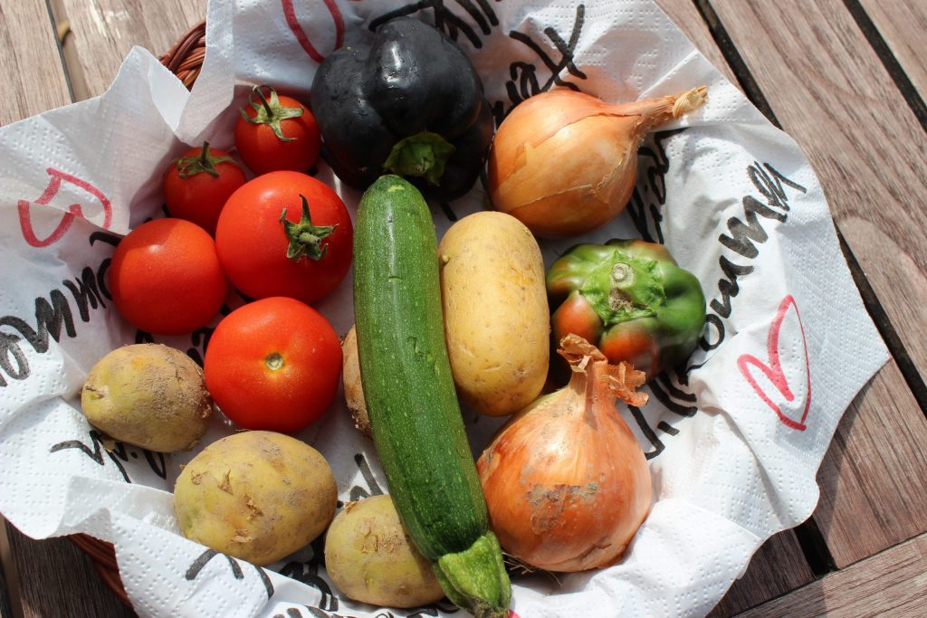 Fresh vegetables including tomatoes, potatoes, onions, courgette, and peppers arranged in a basket on a wooden table.