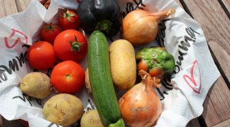 Fresh vegetables including tomatoes, potatoes, onions, courgette, and peppers arranged in a basket on a wooden table.
