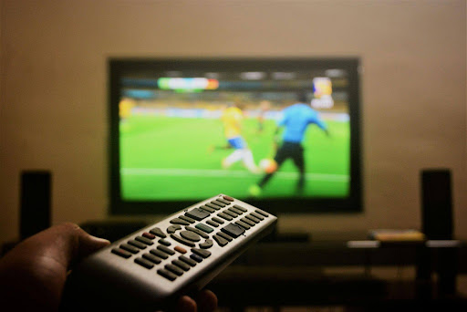 Person holding a TV remote while watching a football match on a television, representing sports entertainment and digital audience engagement.