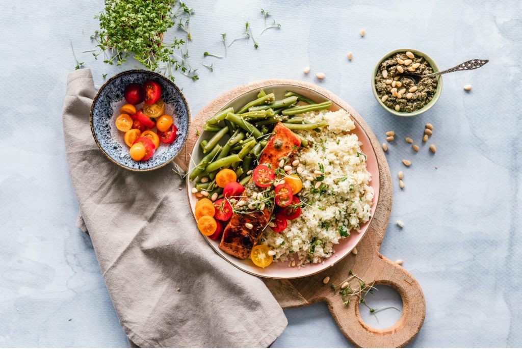 A balanced meal with grilled salmon, couscous, green beans, cherry tomatoes, pesto and pine nuts on a wooden board.