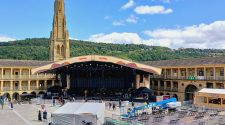 Outdoor stage set up in a historic Yorkshire courtyard with a church tower and green hillside in the background.
