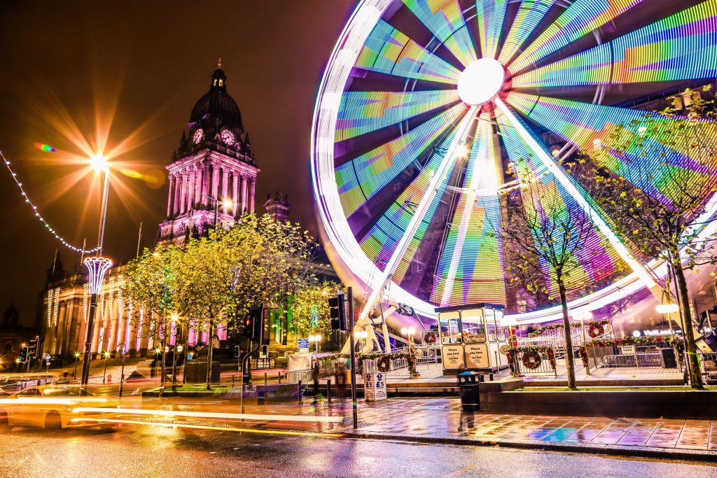 Leeds city centre at night with a brightly lit Ferris wheel spinning beside Leeds Town Hall, colourful lights reflecting on the wet pavement.