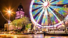 Leeds city centre at night with a brightly lit Ferris wheel spinning beside Leeds Town Hall, colourful lights reflecting on the wet pavement.