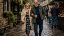 Stylish couple in smart-casual layered outfits walking along a damp cobbled street outside a countryside gastropub and outdoor food market.