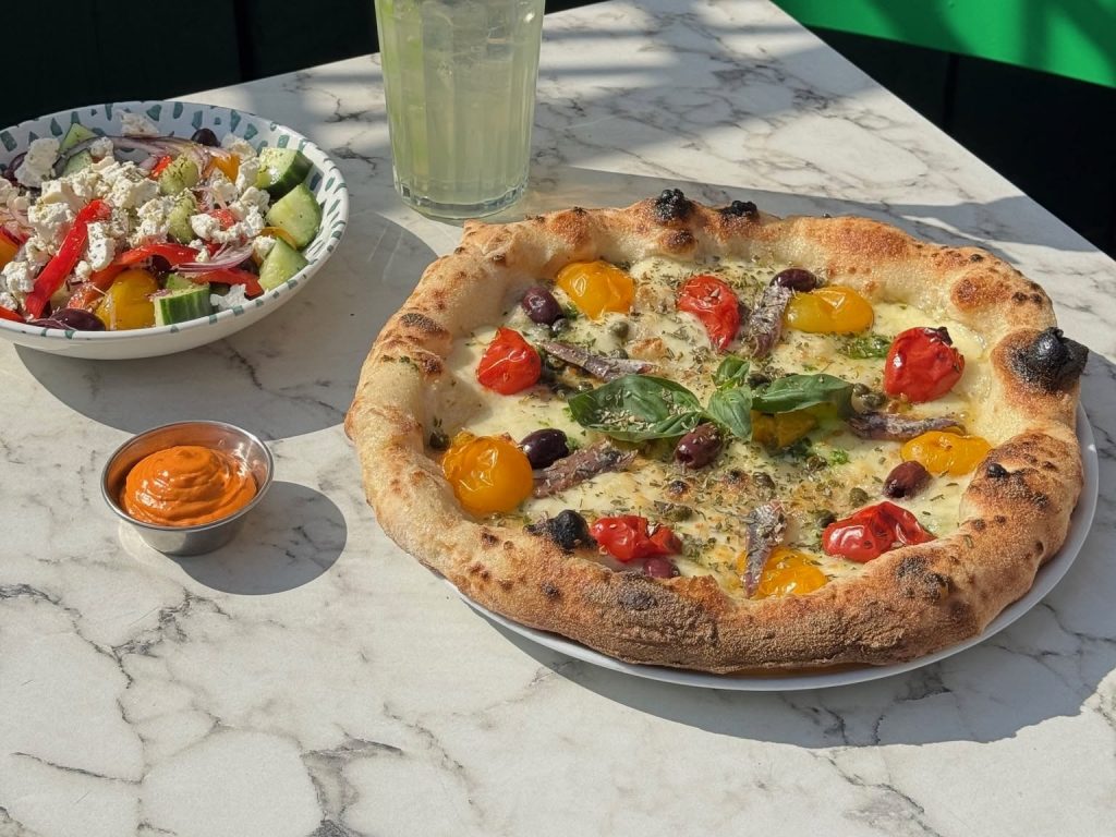 Neapolitan-style pizza topped with tomatoes, olives, anchovies and basil, served with Greek salad and chilli dip on a marble table.