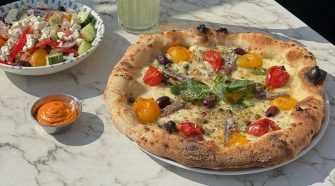 Neapolitan-style pizza topped with tomatoes, olives, anchovies and basil, served with Greek salad and chilli dip on a marble table.