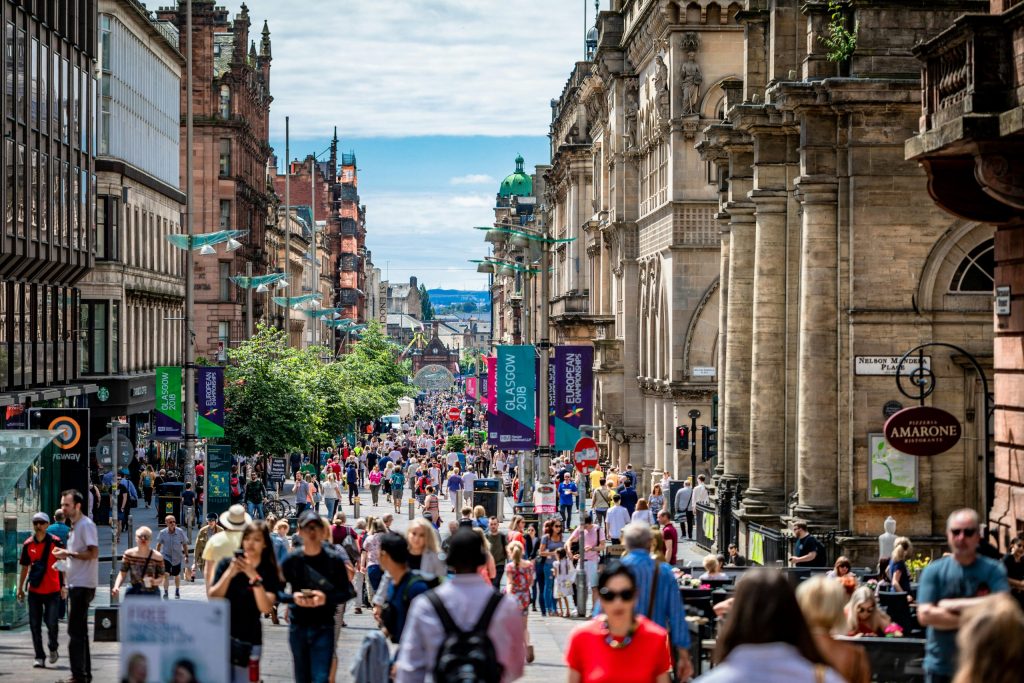 Busy pedestrianised Glasgow city-centre street lined with historic buildings, shops, restaurants and outdoor seating on a sunny day.