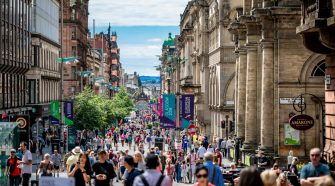 Busy pedestrianised Glasgow city-centre street lined with historic buildings, shops, restaurants and outdoor seating on a sunny day.
