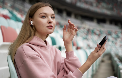 Young woman sitting in a stadium wearing wireless earbuds and holding a smartphone while following mobile sports content.