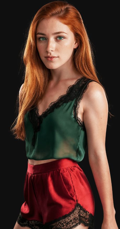 Portrait of a red-haired woman with green eyes standing against a black background, wearing a dark green lace-trim camisole and red satin shorts with black lace detailing.