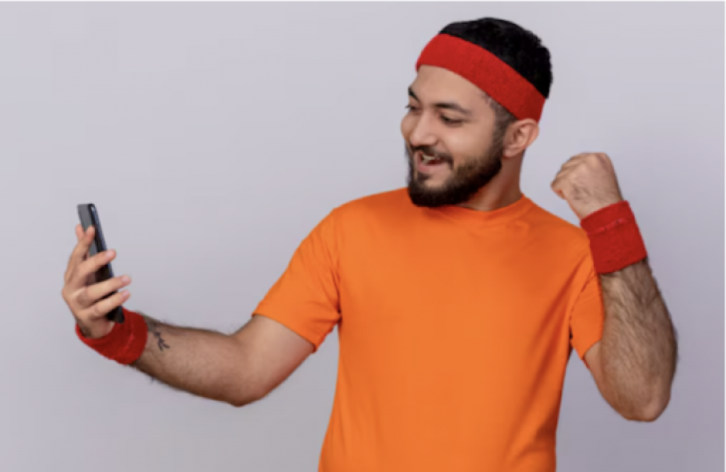 Male sports fan in orange activewear celebrating while checking live football updates on his smartphone.