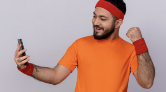 Male sports fan in orange activewear celebrating while checking live football updates on his smartphone.