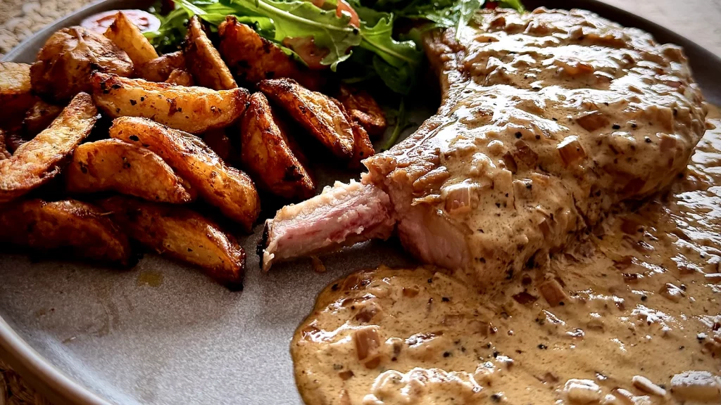 Juicy pork chop covered in creamy peppercorn sauce with crispy potatoes and rocket salad