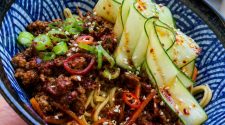 Sticky Korean-style beef mince noodles topped with chilli-oil cucumber ribbons, spring onions and sesame seeds in a blue patterned bowl.