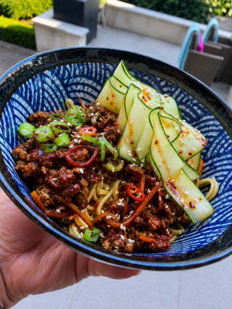 Sticky Korean-style beef mince noodles topped with chilli-oil cucumber ribbons, spring onions and sesame seeds in a blue patterned bowl.