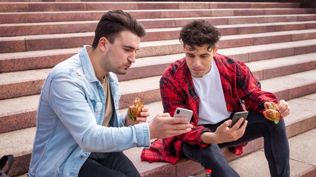 Two young men sitting on outdoor steps eating burgers while checking football updates on their smartphones.