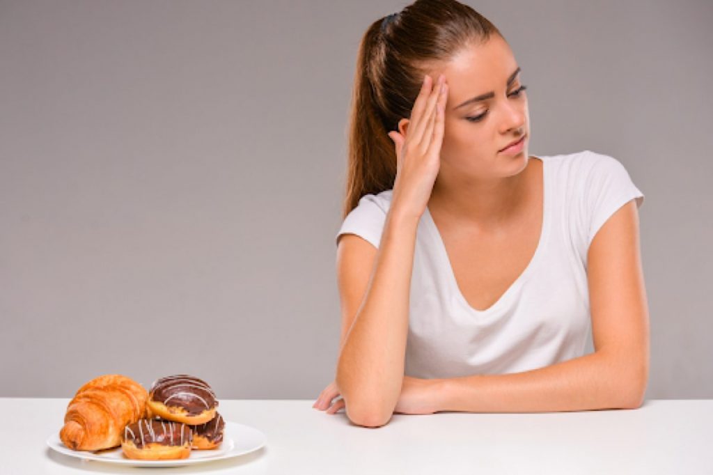 Woman in a white t-shirt sitting at a table, looking thoughtfully at a plate of pastries including donuts and a croissant.