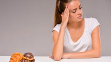 Woman in a white t-shirt sitting at a table, looking thoughtfully at a plate of pastries including donuts and a croissant.