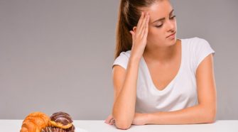 Woman in a white t-shirt sitting at a table, looking thoughtfully at a plate of pastries including donuts and a croissant.
