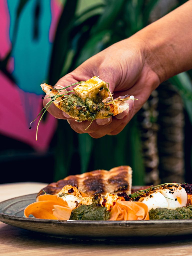 Flatbread topped with green sauce and cheese held above poached eggs and carrot ribbons at House of Koko Leeds