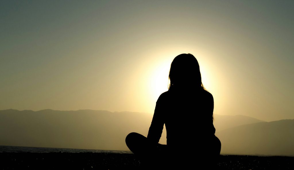 Silhouette of a woman sitting cross-legged on a beach at sunset, with mountains in the distance