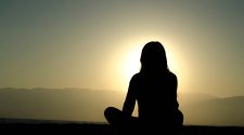 Silhouette of a woman sitting cross-legged on a beach at sunset, with mountains in the distance