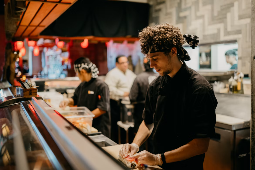 Chef preparing sushi at a counter with colleagues in a commercial kitchen