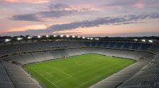An empty floodlit football stadium at dusk, with a green pitch and grey and blue seats under a soft evening sky.